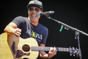 Male guitarist wearing a cap and sunglasses, singing into a microphone while playing an acoustic guitar on stage.