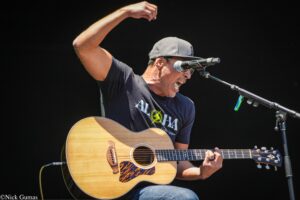 Male guitarist performing on stage with an acoustic guitar, wearing a gray cap and sunglasses, singing into a microphone to the right.