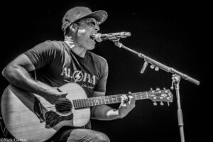 Male musician wearing a cap and sunglasses, singing into a microphone while playing an acoustic guitar on stage.