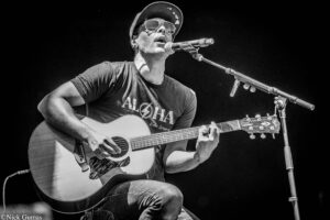 Male musician wearing a cap and sunglasses, singing into a microphone while playing an acoustic guitar on stage (black-and-white photo).