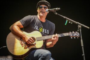 Male guitarist wearing sunglasses and a cap, playing an acoustic guitar into a microphone on stage