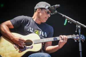 Musician wearing a cap and sunglasses playing an acoustic guitar on stage with a microphone nearby.