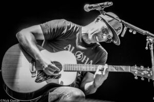 Musician in a hat and sunglasses playing an acoustic guitar on stage with a microphone above him (black-and-white photo).