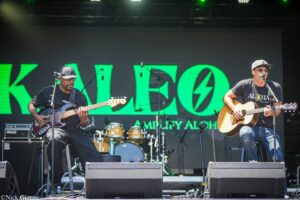 Two male musicians on a stage, one playing bass and the other acoustic guitar, with a green neon backdrop and drums behind them at an outdoor concert.