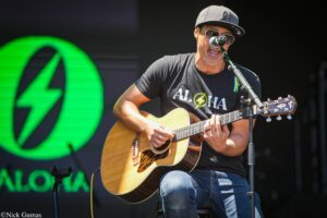 Male guitarist performing on stage, wearing a cap and sunglasses, singing into a microphone while playing an acoustic guitar on stage.