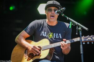 Male musician wearing a black cap and sunglasses plays an acoustic guitar on stage, singing into a microphone.