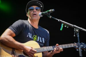 Male musician wearing a cap and sunglasses playing an acoustic guitar on stage during a performance.