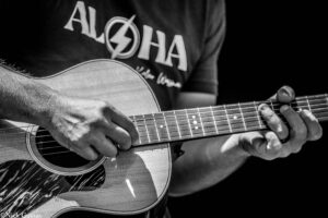 Close-up of hands playing an acoustic guitar, strings and soundhole visible in black-and-white.