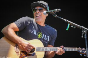 Guitarist performing on stage, wearing a black cap and aviator sunglasses, playing an acoustic guitar into a microphone.