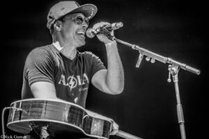 Male musician singing into a microphone while playing an acoustic guitar on stage; wearing a cap and sunglasses (black-and-white).