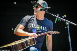 Male guitarist wearing a cap and sunglasses performs on stage, playing an acoustic guitar with a microphone nearby.