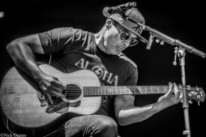 Musician wearing sunglasses and a cap, strumming an acoustic guitar on stage with a microphone stand nearby (black and white).