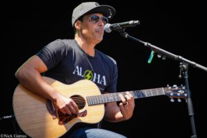 Male musician wearing a cap and sunglasses, singing into a microphone while playing an acoustic guitar on stage.