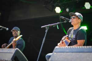 Two musicians perform on stage, one singing into a microphone while playing guitar, wearing a cap and sunglasses; green stage lights in the background.