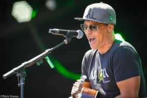 Male musician singing into a microphone while playing a guitar on stage, wearing a gray cap and sunglasses.