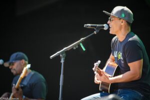 Two guitarists performing on a dimly lit stage, one singing into a microphone while wearing a cap and sunglasses.