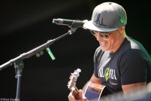 Male guitarist wearing sunglasses and a gray cap, singing into a microphone on stage while playing an acoustic guitar