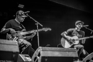 Two musicians on stage: left is a bass guitarist in a cap; right is a guitarist/singer with sunglasses singing into a microphone.