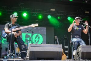 Two male musicians perform on a stage under green lights; guitarist left wearing cap and black shirt, singer right in sunglasses and cap with a microphone.