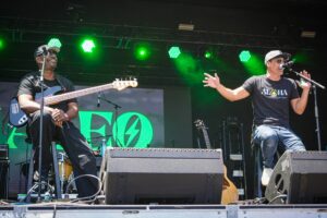 Two male musicians perform on a stage under bright green lights: a bass guitarist on the left and a vocalist with a microphone on the right, both wearing dark shirts and caps.