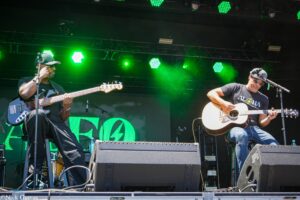 Two musicians play guitars on a lit outdoor stage with bright green lights and equipment in the background.