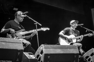 Two musicians performing on stage: one playing a bass guitar and the other singing and playing an acoustic guitar into a microphone.