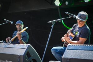 Two guitarists perform on a dark stage: a man in a gray cap and sunglasses on the right, playing an acoustic guitar, with a keyboardist in a black cap on the left.