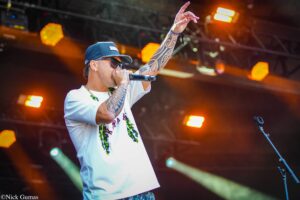 Male singer on stage wearing a blue cap and sunglasses, microphone in hand, tattooed arm raised as orange stage lights glow behind him