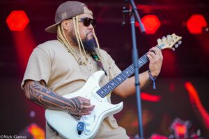 Guitarist wearing a backward cap and sunglasses, with long braids, playing a white electric guitar on stage.