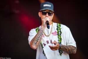 Male performer wearing a Hawaii cap, sunglasses, and a white shirt with a tropical lei, singing into a microphone on stage with tattooed arms visible.