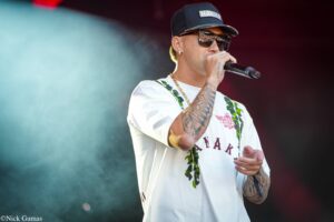 Male singer wearing a cap and sunglasses, holding a microphone on stage; tattoos visible on his arm.