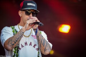 Male performer wearing a navy cap that says 'HAWAII' and dark sunglasses, singing into a microphone on a stage with red lighting in the background.