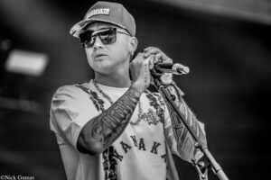 Male vocalist on stage wearing sunglasses and a cap, gripping a microphone, showcasing full arm tattoos in black-and-white.