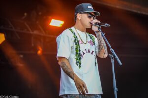 Male singer performing on a stage, wearing a cap, sunglasses, white graphic shirt, gold chain, and visible tattoos while singing into a mic under orange stage lights.