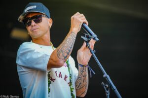 Male singer with tattoos and a blue Hawaii cap, wearing sunglasses, singing into a handheld mic on stage.
