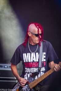 Bass guitarist with bright red dreadlocks performing on a dim stage, wearing sunglasses and a black graphic T-shirt with a checkered strap.