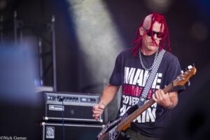 Bassist with red dreadlocks wearing sunglasses plays a bass on stage with a checkered strap.