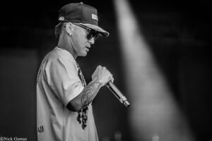 Side profile of a male singer wearing a cap and sunglasses, holding a microphone on stage (black and white).