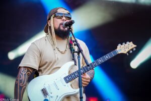 Male guitarist performing on stage, wearing sunglasses, a backward cap, and gold chains, playing a white electric guitar into a microphone while singing/performing.