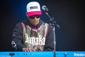 Male keyboardist in camouflage jersey, black hoodie, white cap with pink brim and sunglasses, playing a blue Yamaha keyboard with a microphone nearby on stage.