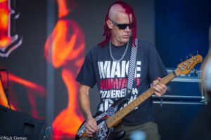 Male bassist with red dreadlocks and sunglasses playing a bass on stage, wearing a dark graphic t-shirt and a checkered strap.