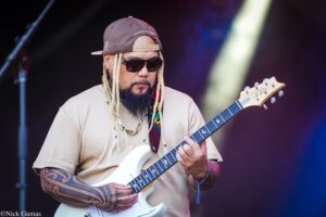 Guitarist playing a white electric guitar on stage, wearing a brown cap, sunglasses, and braided hair with tattoos showing on his arms.