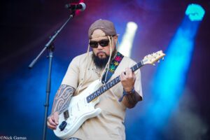Guitarist wearing sunglasses and a backward cap plays a white electric guitar on stage.