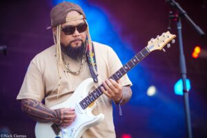 Guitarist with long braided hair, cap and sunglasses playing a white electric guitar on stage with blue lighting behind him.