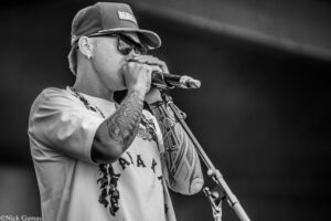 Male singer performing into microphone, wearing cap, sunglasses, and heavily tattooed arms in a black-and-white photo