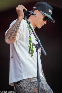 Male singer performing on stage, gripping a microphone stand with a tattooed forearm visible, wearing a cap, sunglasses, a white t‑shirt, and camo shorts, microphone and mic stand in front of him.