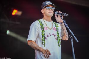 Male singer on stage wearing a cap, mirrored sunglasses, and a green lei, holding a microphone.
