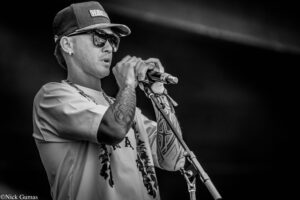 Male singer on stage wearing cap and sunglasses, gripping a microphone with tattooed arms visible, performing intently.