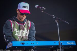 Musician playing a bright blue Yamaha keyboard on stage, wearing a camouflage jersey, black hoodie, pink-and-white cap, and dark sunglasses, with a microphone on a stand nearby