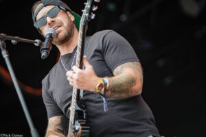 Male guitarist wearing sunglasses and a cap, singing into a microphone on stage